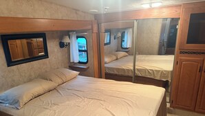 A bedroom with two beds, a window with curtains, a mirror, and wooden cabinets. - Fantastic RV with AC in Muskegon (Muskegon)