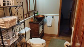 Een badkamer met toilet, een wastafel met een waskom, een spiegel met verlichting, een handdoekhouder en een plank met manden.