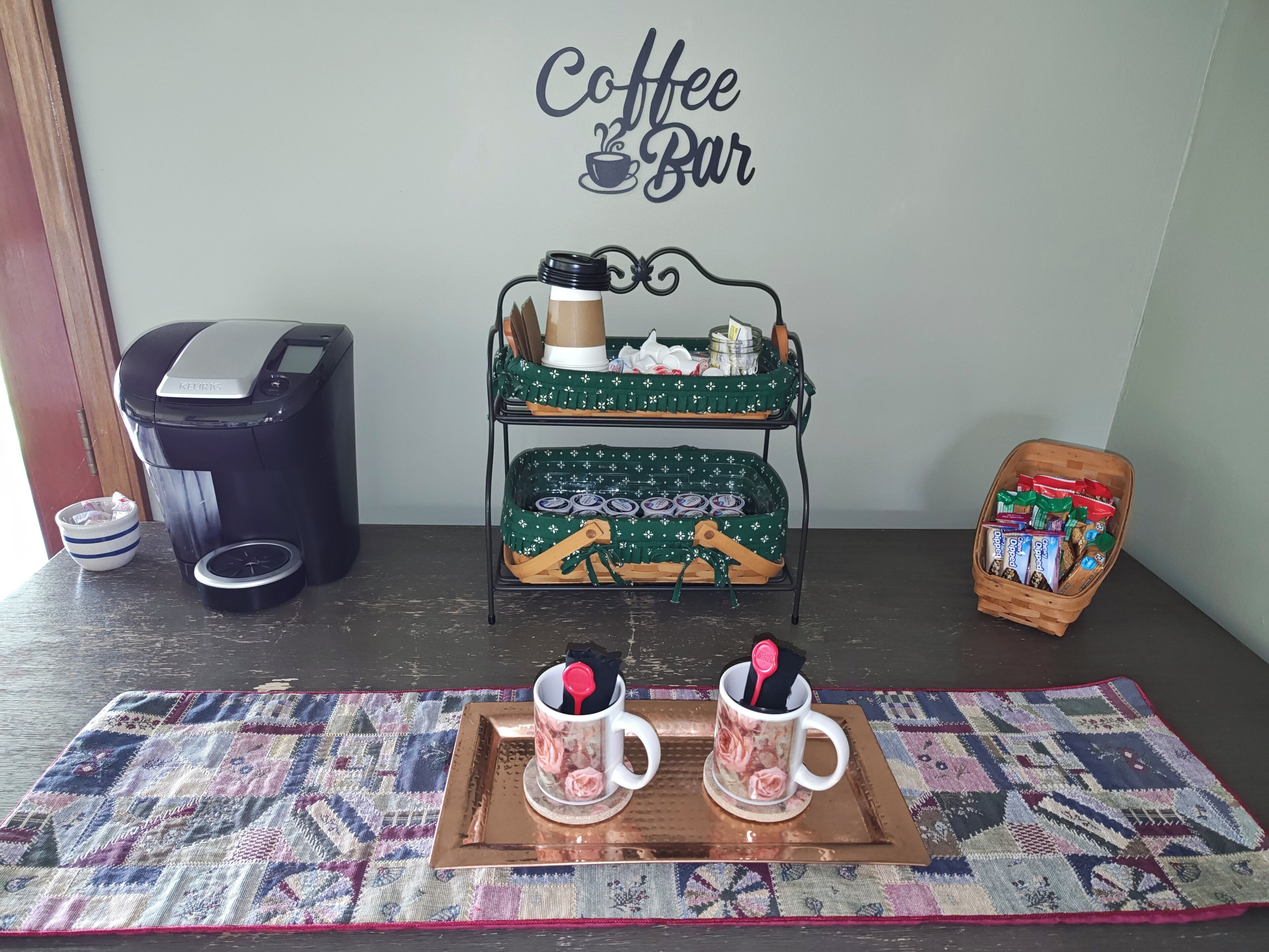 Een koffiebar-opstelling met een Keurig koffiezetapparaat, een dienblad met twee mokken, een koffiehoekje met een kop en schotel, en een mandje met snacks.