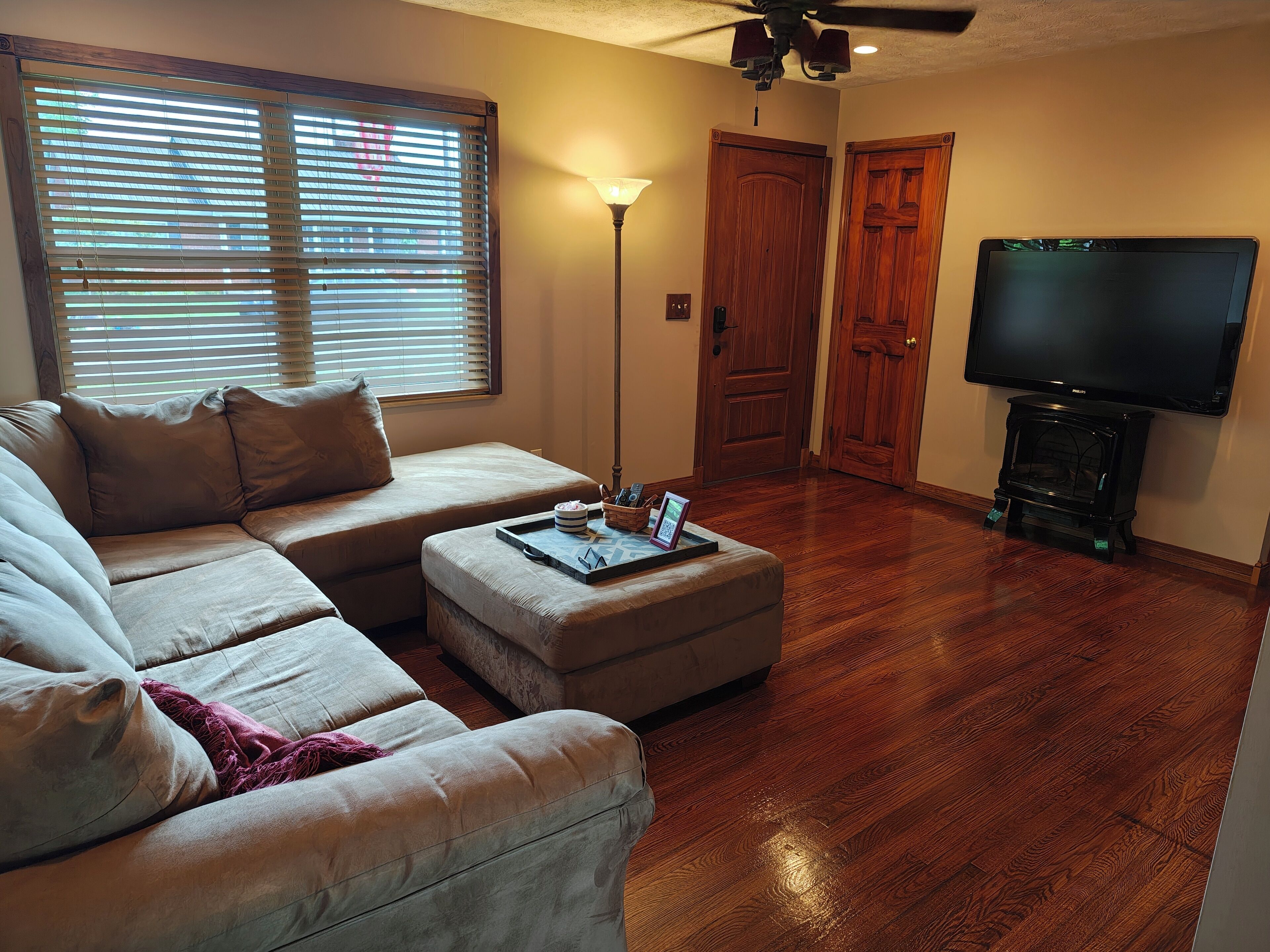 A living room with a sectional sofa, ottoman, wooden flooring, a television, and a fireplace.