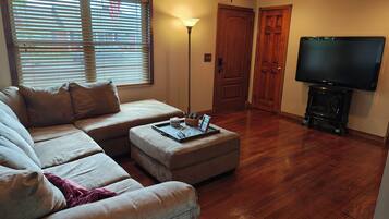 A living room with a sectional sofa, ottoman, wooden flooring, a television, and a fireplace.