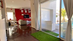 Een moderne woonkamer met een rode accentmuur, een flatscreen televisie, een eethoek met een witte tafel en stoelen, en een schuifpui die uitkomt op een balkon met uitzicht op een zwembad.