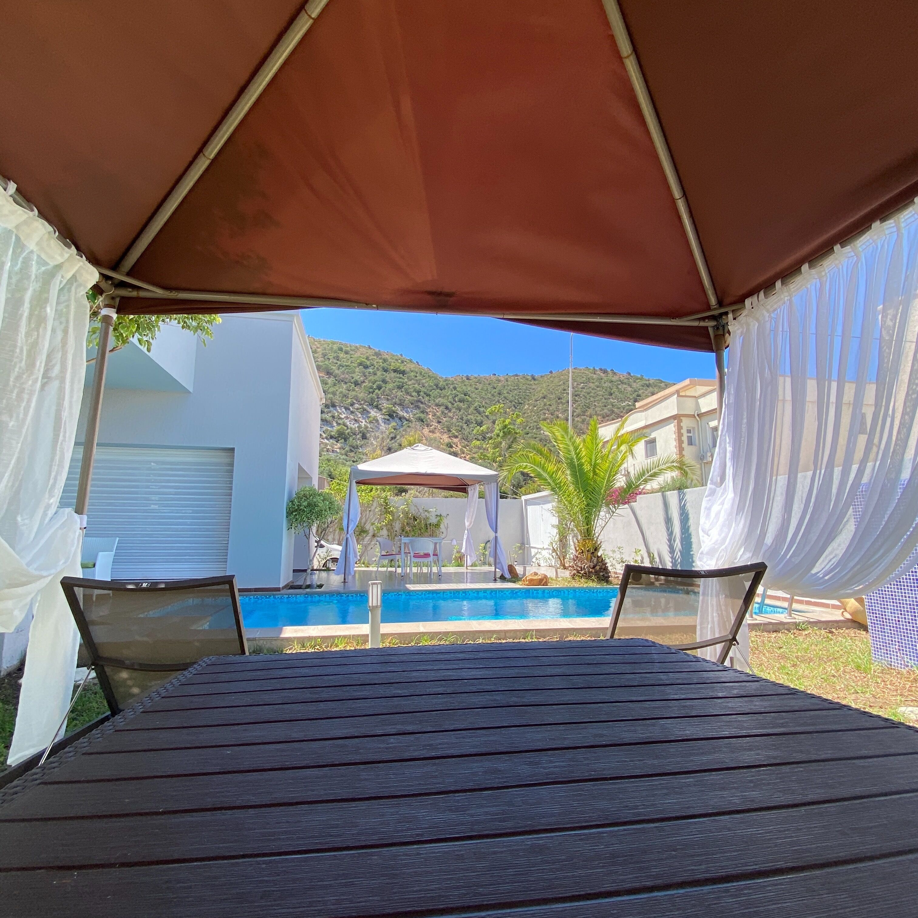 A covered outdoor seating area with a pool, white buildings, and a hill in the background.