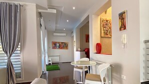 Een moderne woonkamer met een witte bank, een eettafel met twee stoelen, en een salontafel met een glazen blad. Er is een radiator, een witte telefoon, en kunstwerken aan de muur.