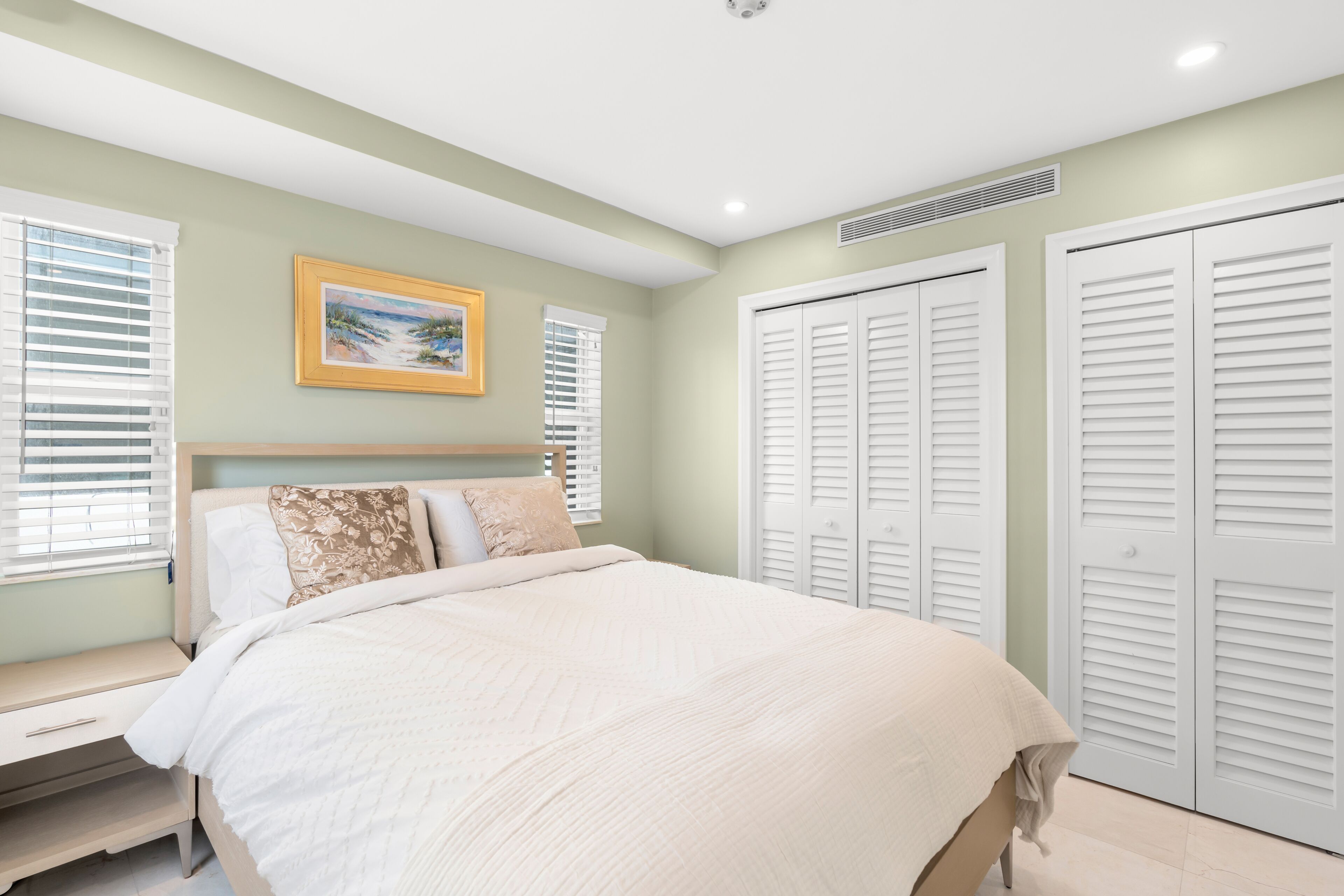 A bedroom with a bed, a bench, a painting, and white closets.