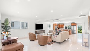 Ein geräumiges Wohnzimmer mit einer Couch, Sesseln, einem Couchtisch und einem Fernseher. Im Hintergrund ist eine Küche zu sehen.
