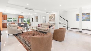 Ein großzügiger Wohnbereich mit einer Couch, Sesseln und einem Couchtisch. Im Hintergrund ist eine Küche zu sehen, und eine Treppe führt zu einer anderen Etage.