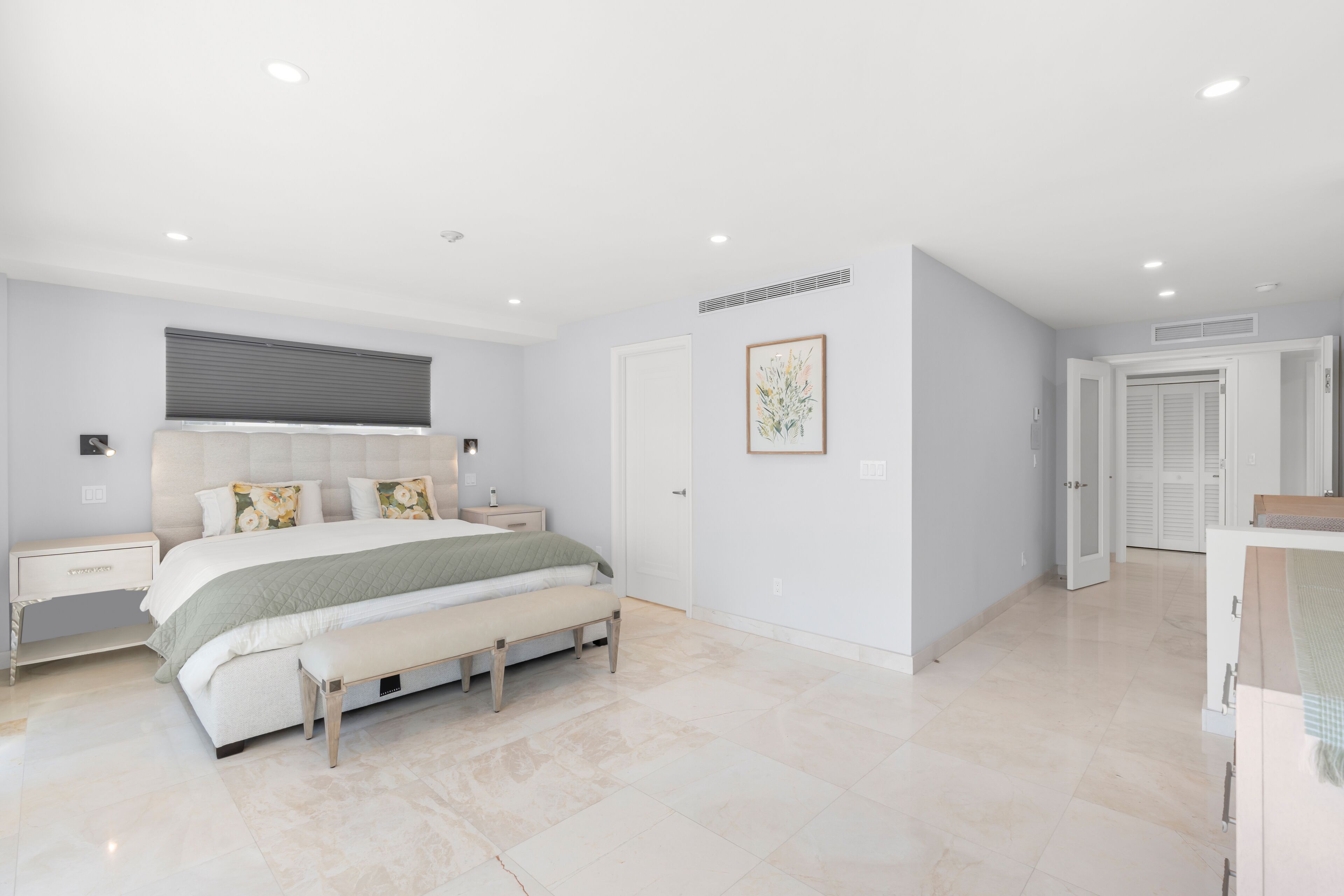 A modern bedroom with a large bed, bedside tables, a bench, and a door leading to another room.