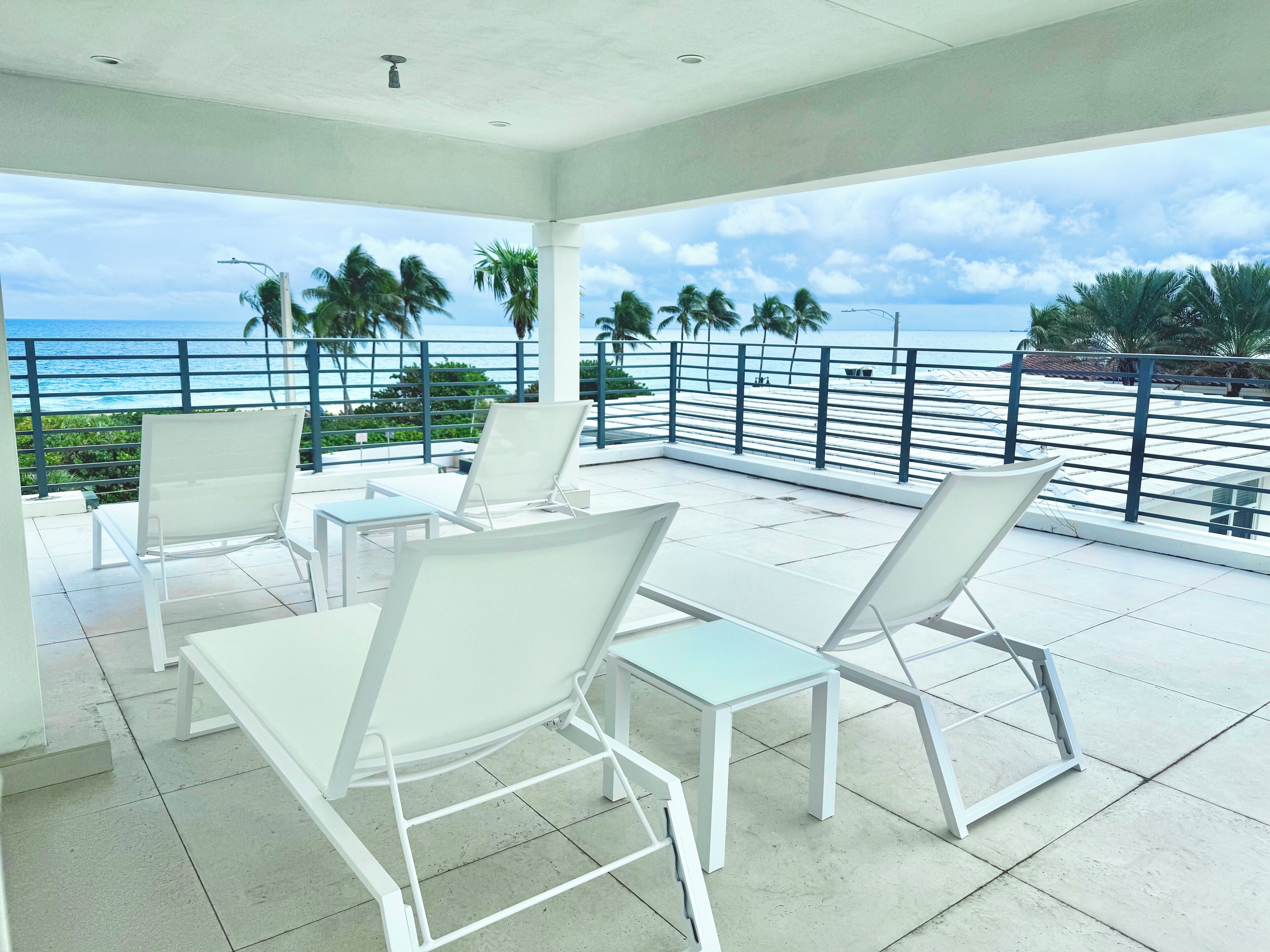 A balcony with white chairs and tables, overlooking a beach with palm trees and the ocean.