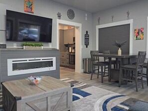 A modern living room with a fireplace, a dining area with a table and chairs, a kitchen in the background, and a TV mounted on the wall. - CASITA, 1 bedroom, living room, kitchen, bathroom, Wi-Fi, parking, close to NNU. (Nampa)