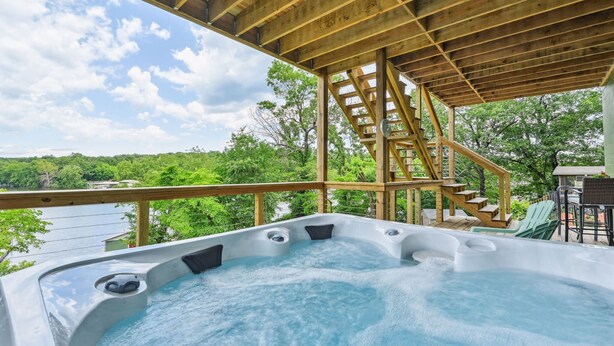 A hot tub area with a wooden deck, a staircase, and a view of a lake surrounded by trees.