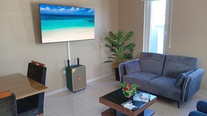 A living room with a large flat-screen TV, a grey sofa, a wooden coffee table with remote controls, a small table with chairs, and a potted plant. - Island Escape Home in Noord, Aruba (Noord)