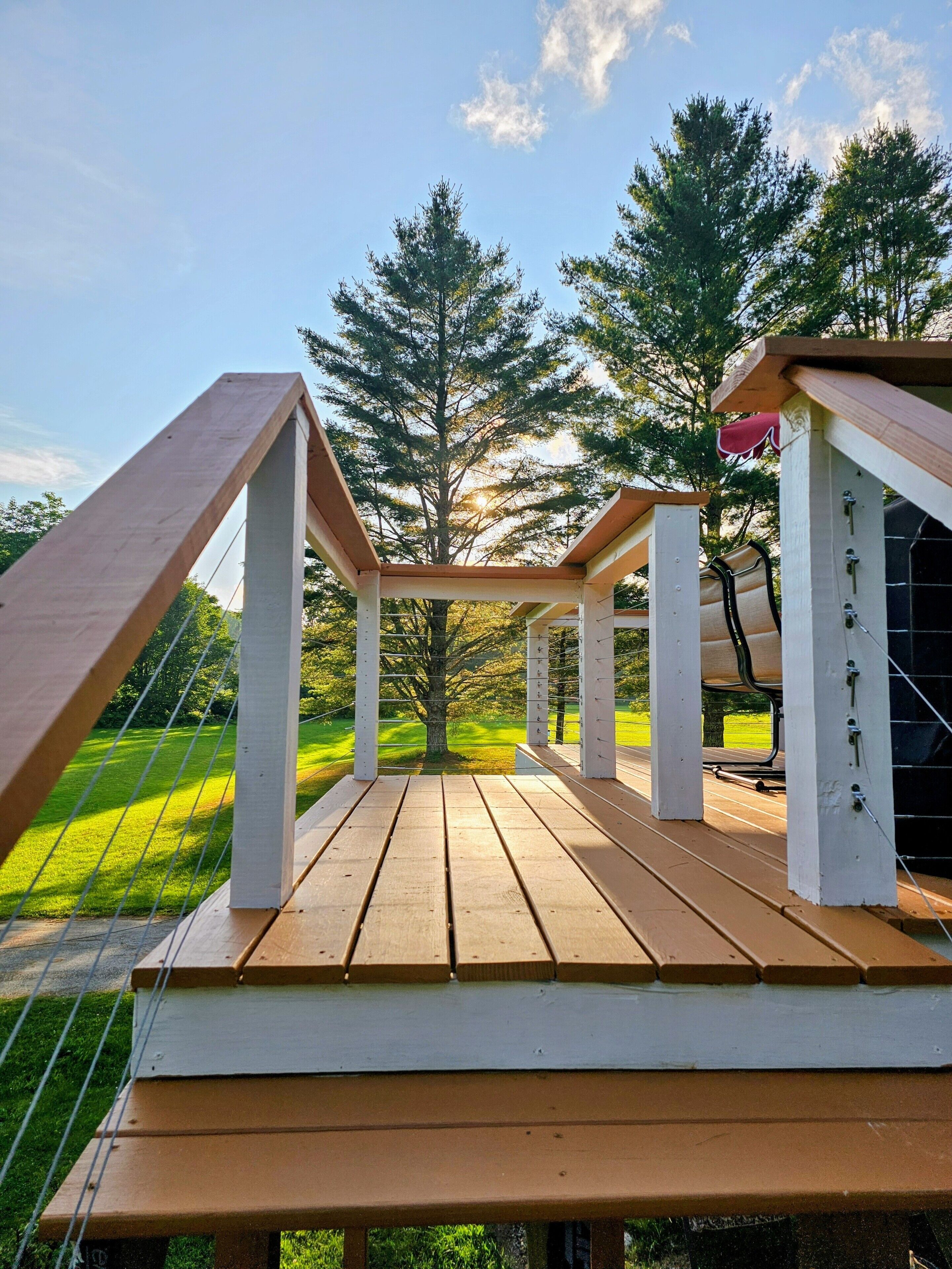 A wooden deck with a pergola structure, surrounded by trees and a grassy area.