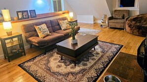 A cozy living room with a brown leather sofa, a dark coffee table, and a patterned rug. There are two side tables with lamps, a bookshelf, and a window with blinds. - Cozy cabin >> Hottub, WiFi, AC. Easy access to outdoors activities year around (Hancock)