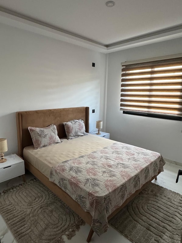 Une chambre à coucher comprenant un lit, deux tables de chevet, une commode, une fenêtre avec des stores et un tapis à motifs.