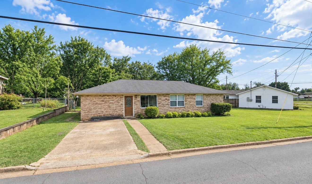 A single-story brick house with a front yard, a driveway, and a small white building in the background.