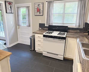 A kitchen with a white stove, dark tiled floor, and a window with curtains. - The Little House Near UA & Downtown (Tuscaloosa)