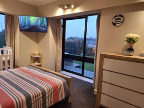 Een slaapkamer met een bed, een televisie, een digitale klok, een raam met uitzicht, en een tafeltje met een bloemenvaas.
