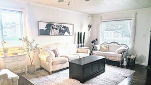 A living room with a white sofa, a black coffee table, and a patterned rug. There are two windows with blinds, a ceiling fan, and a framed picture on the wall. - Teton Heritage House | Yellowstone Central Hub | Magical Tree (Teton)