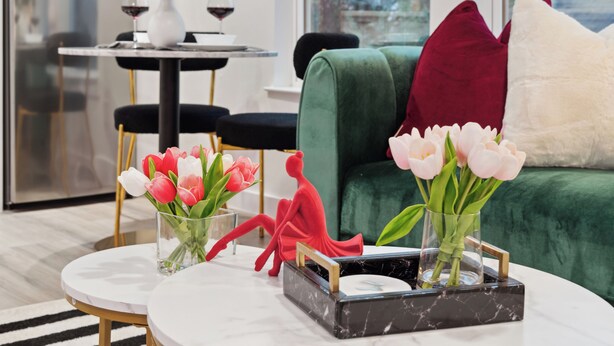 Ein modernes Wohnzimmer mit einem runden Tisch, zwei Vasen mit Blumen, einem grünen Sofa und einem rot-weißen Kissen.