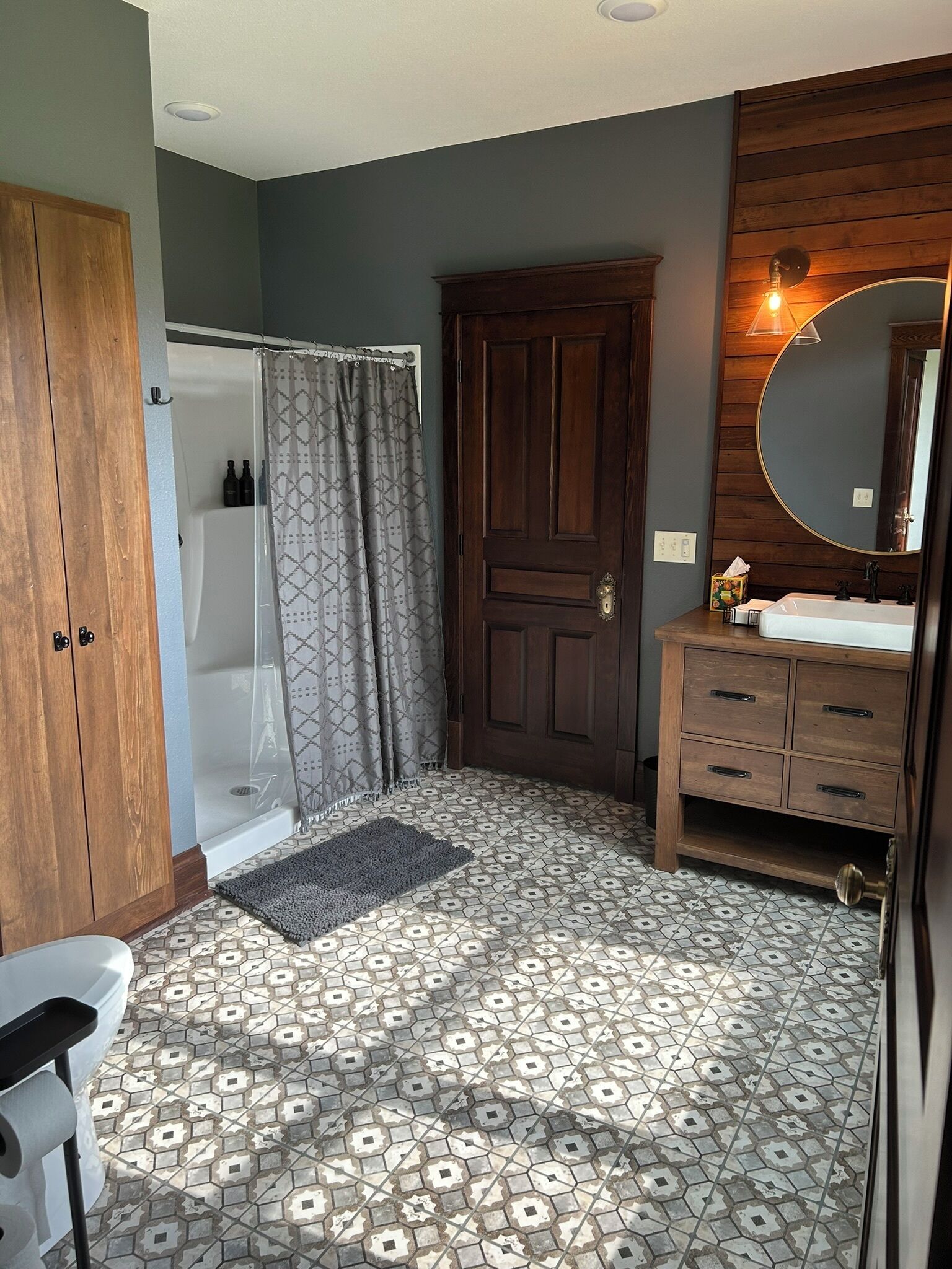 A bathroom with a shower, a wooden door, a vanity with a mirror, and patterned floor tiles.