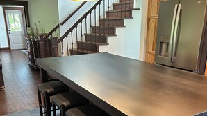 A kitchen with a long island, bar stools, a stainless steel refrigerator, and a staircase with wooden steps. - A Peaceful Driftless Farm Stay Awaits Near A Trout Stream (Muscoda)