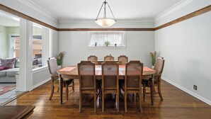 Een eetruimte met een ronde tafel, zes houten stoelen, een hanglamp en een raam met planten.