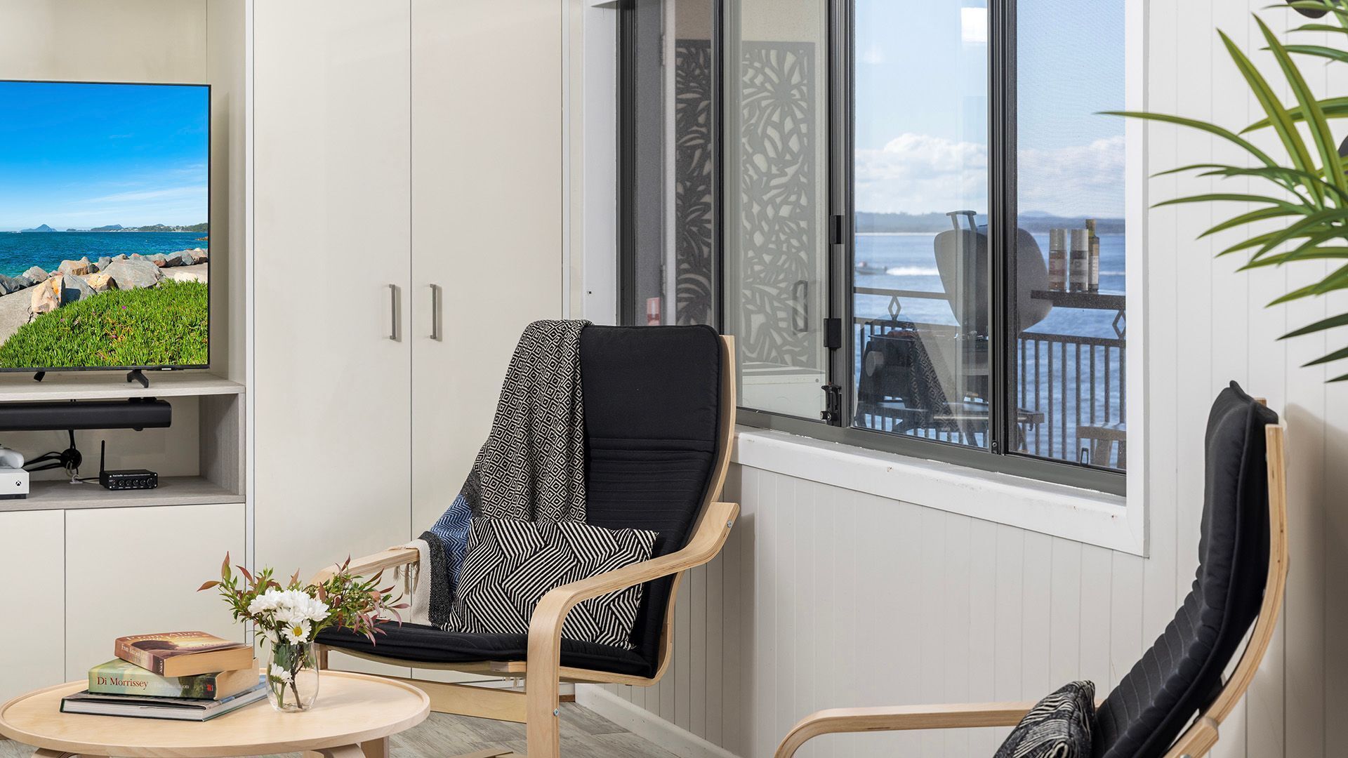 Una sala moderna con vistas al mar, un sillón cómodo, una mesita con libros y flores, y un televisor de pantalla plana sobre un mueble.