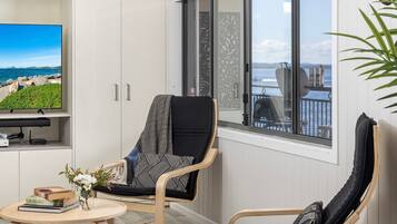 Una sala moderna con vistas al mar, un sillón cómodo, una mesita con libros y flores, y un televisor de pantalla plana sobre un mueble.