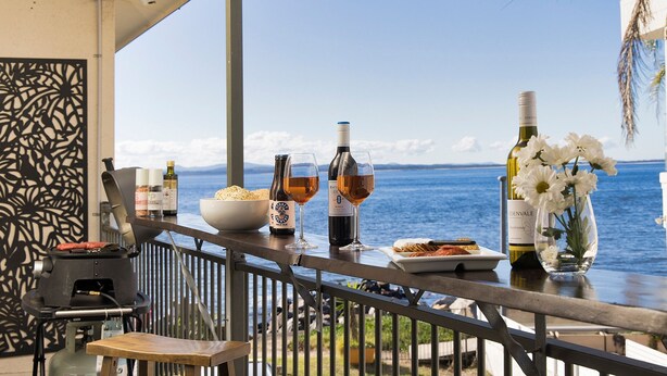 A balcony with a view of the sea, featuring a grill, wine bottles, glasses, and a bowl of bread.