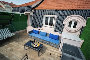 A rooftop terrace with a wicker sofa, a dining table with a tray of food, and a green lawn.