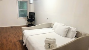 A single-bed room with a wooden floor, a window with blinds, a black chair, and a bedside table.