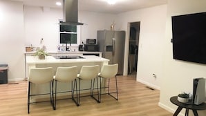 A modern kitchen with a central island, bar stools, a large refrigerator, and a flat-screen TV on a stand.