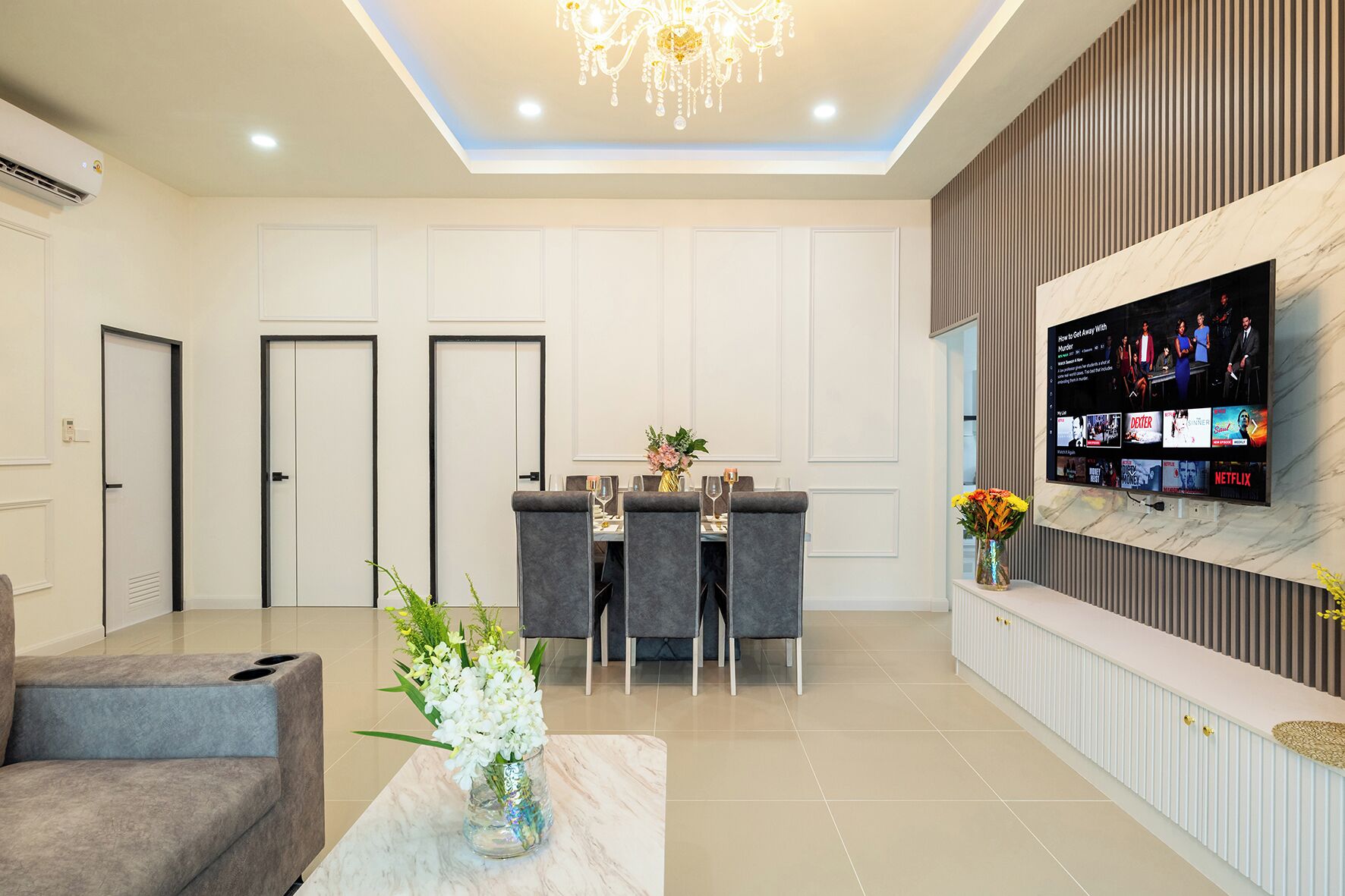 A modern living room with a dining area, a sofa, a TV mounted on the wall, and a chandelier.