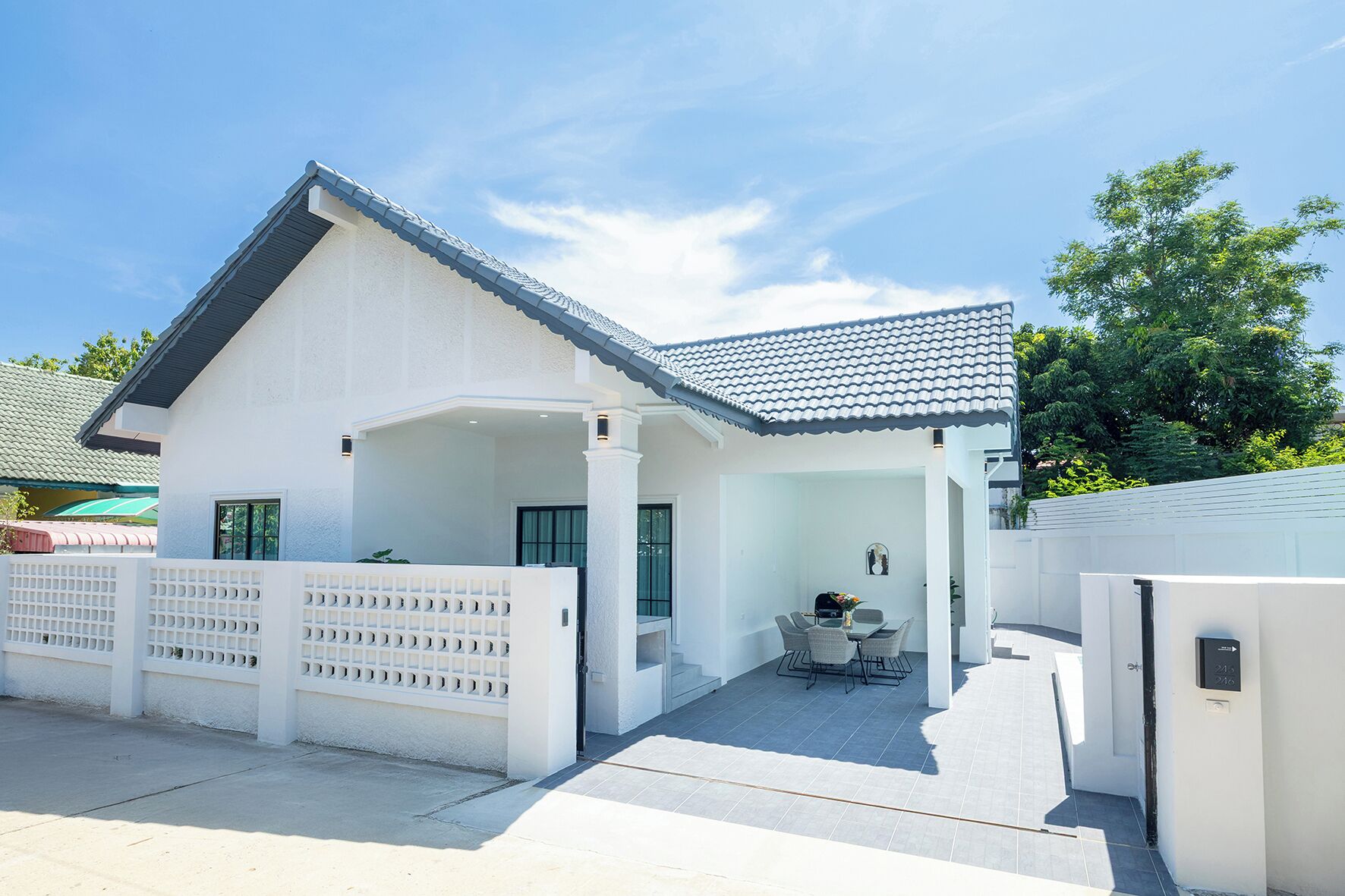 A white house with a tiled roof, a covered patio with a table and chairs, and a white fence.