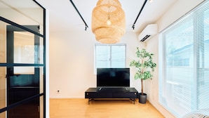 A modern living room with a flat-screen TV, a woven light fixture, a potted plant, and large windows with blinds.