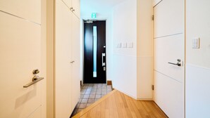 A narrow hallway with white walls and wooden flooring, leading to a door with a black frame and a light switch above it.