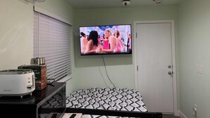 Una stanza con televisione a parete, ventilatore a soffitto, finestra con persiane e angolo cottura dotato di tostapane e macchina del caffè.