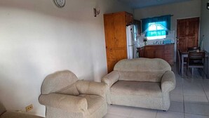 Una sala de estar con dos sofás color beige, un aparador de madera y un área de cocina con refrigerador y ventana.