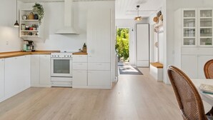 Una cocina moderna con armarios blancos, encimera de madera y horno empotrado. Hay un comedor con sillas de mimbre y una mesa.