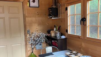 A wooden cabin interior with a dining table, chairs, a clock, framed pictures, and a window with a view of trees.