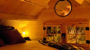 Un dormitorio acogedor con techo de madera, un espejo redondo, una cama con manta estampada y una ventana con vistas a árboles.