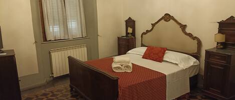 Une chambre avec un sol à carreaux, un lit recouvert d’une couverture rouge, des oreillers blancs et une tête de lit en bois.