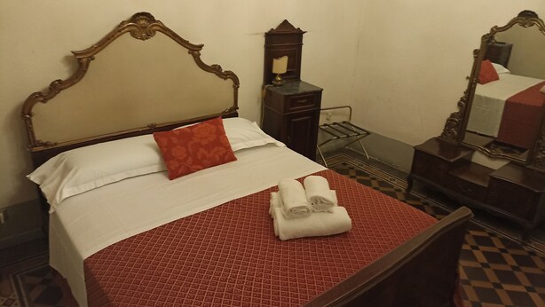 A neatly made bed with a red and white patterned bedspread, a wooden headboard, and a single red pillow. There is a small wooden nightstand with a mirror and a lamp beside the bed.