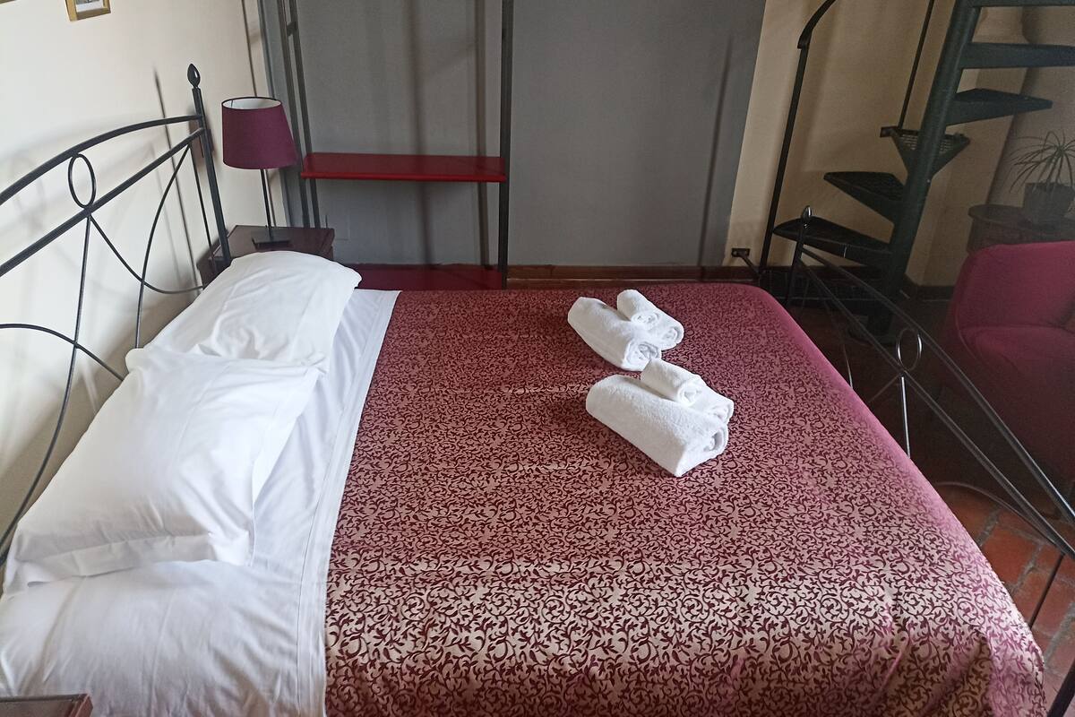 A neatly made bed with white linens and a red patterned bedspread. There are two rolled towels on the bed.