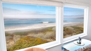 Una habitación con dos ventanas amplias que dan a una playa, una pequeña mesa con un farolillo y una silla.