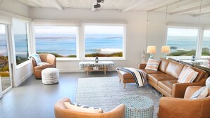 Un salón amplio con un gran ventanal que ofrece vistas al océano. Hay sofás de cuero, un otoman redondos y una mesita de centro.
