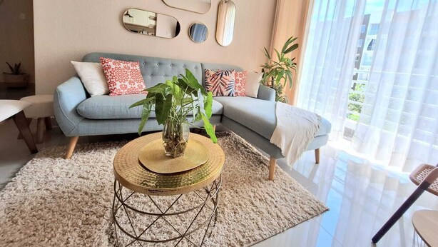 A modern living room with a grey sofa, decorative mirrors, a round coffee table with a plant, and a large window with sheer curtains.
