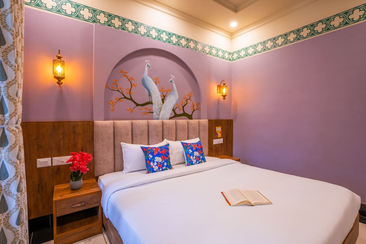 A bedroom with a large bed, two wall-mounted lamps, a decorative peacock mural, and a nightstand with a vase of flowers.