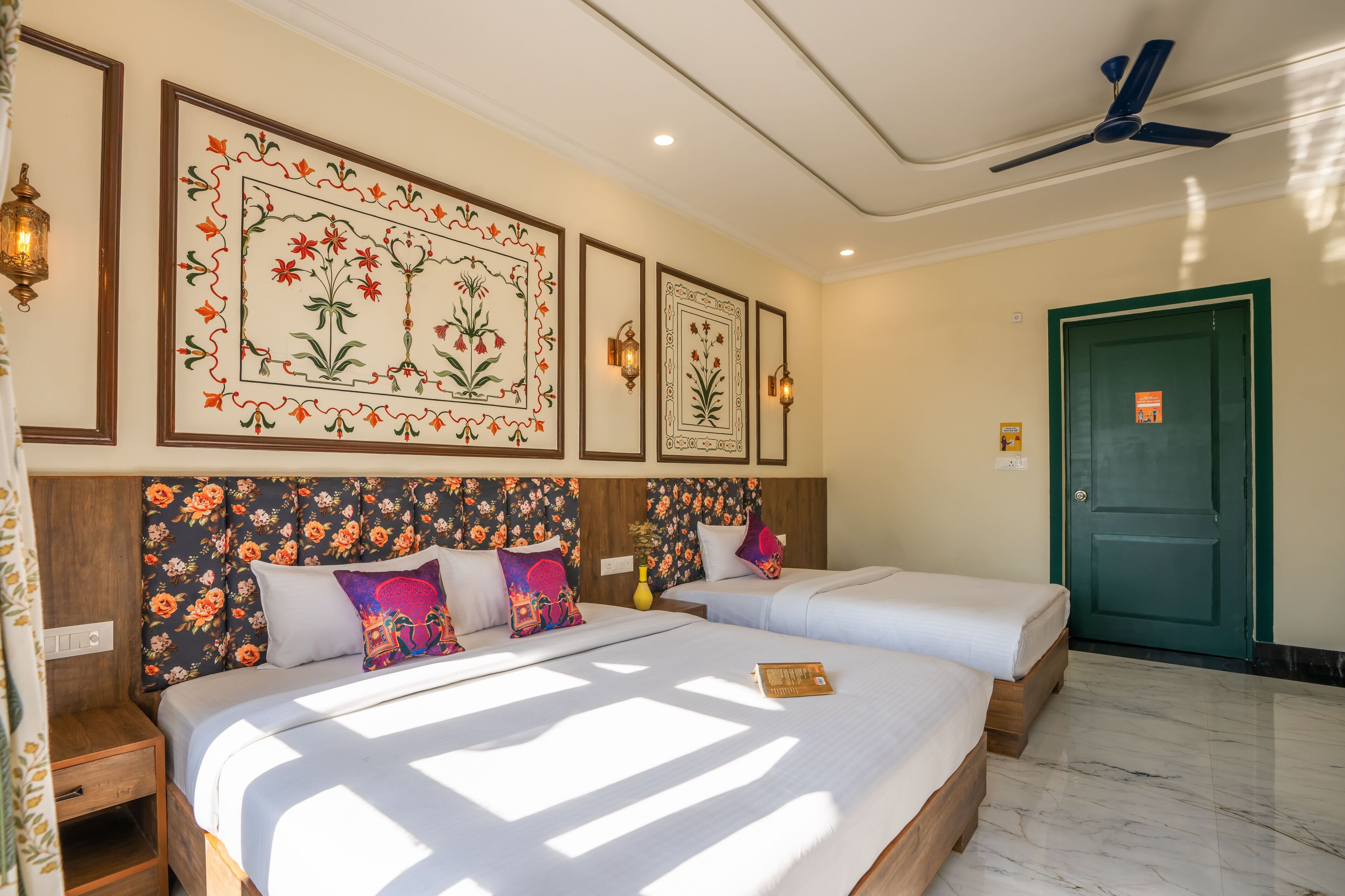 A hotel room with two beds, floral headboards, wall-mounted artwork, and a ceiling fan.